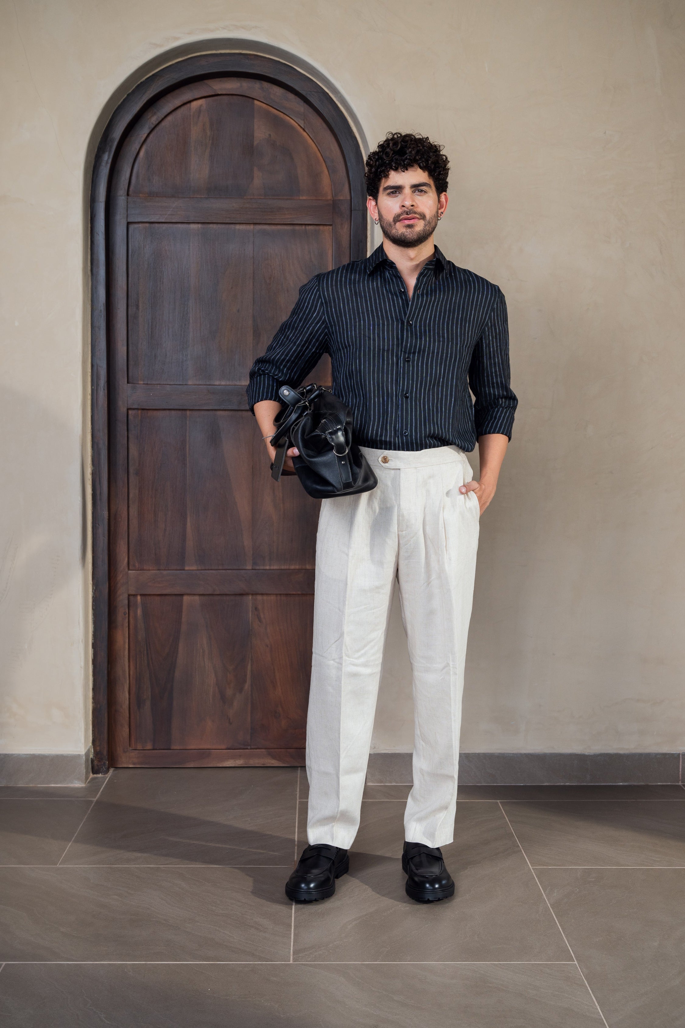 Man in a dark shirt and white pants standing in front of a wooden door.
