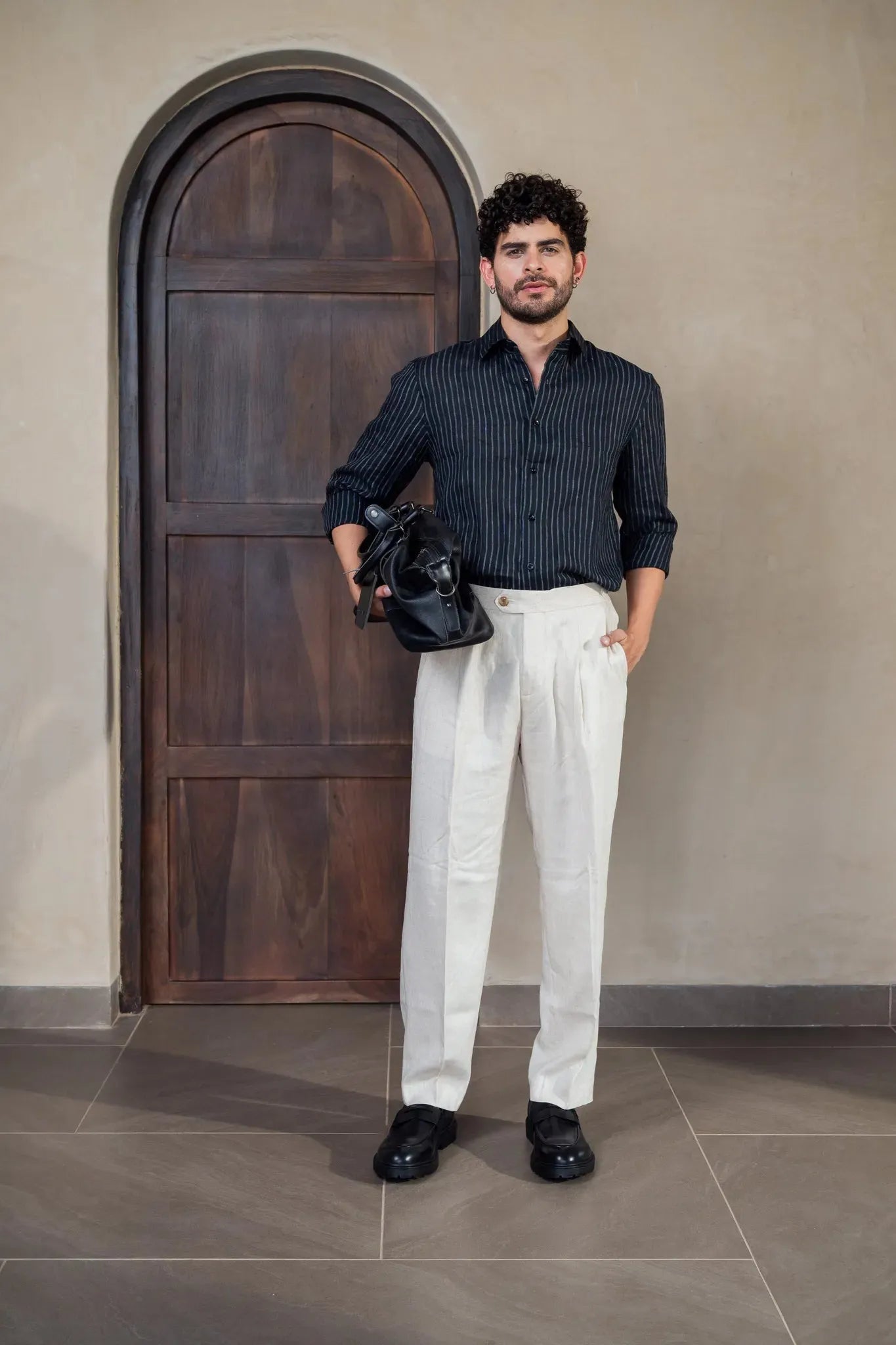Man in a dark shirt and white pants standing in front of a wooden door.